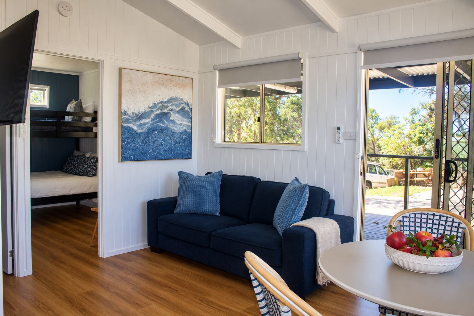 A room with a dark blue sofa, a painting, a window with a view of greenery, and a table with a fruit bowl.
