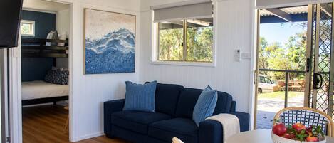 A room with a dark blue sofa, a painting, a window with a view of greenery, and a table with a fruit bowl.