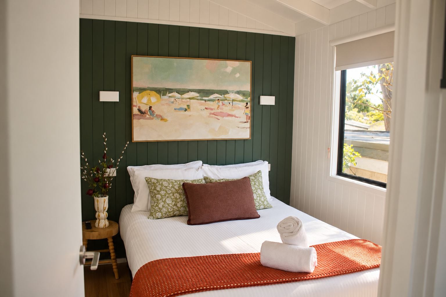 A bedroom with a bed, pillows, a vase with flowers, and a painting on the wall.