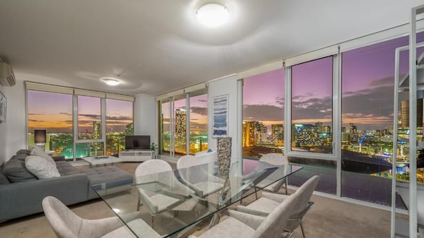 Uma sala de estar moderna com uma mesa de vidro, assentos confortáveis, um sofá e grandes janelas com vista para a cidade ao entardecer.