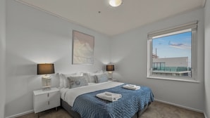 Quarto com cama, duas luminárias de cabeceira, uma janela grande e um quadro na parede.