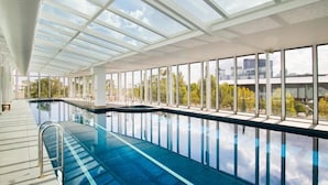 Piscina interna com teto de vidro, janelas amplas e vista para a paisagem urbana.