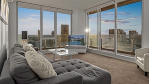 Uma sala de estar ampla com um sofá cinzento, uma mesa de centro e uma grande janela com vista para a cidade.