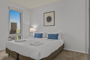 Une chambre d’hôtel avec un grand lit, une fenêtre donnant sur la ville, un tableau encadré au mur et une table de chevet avec une lampe.