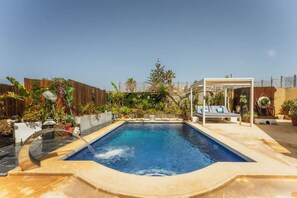 Ein rechteckiger Swimmingpool mit Wasserspiel, umgeben von einem Holzzaun, Topfpflanzen und einer Lounge mit einer Pergola.