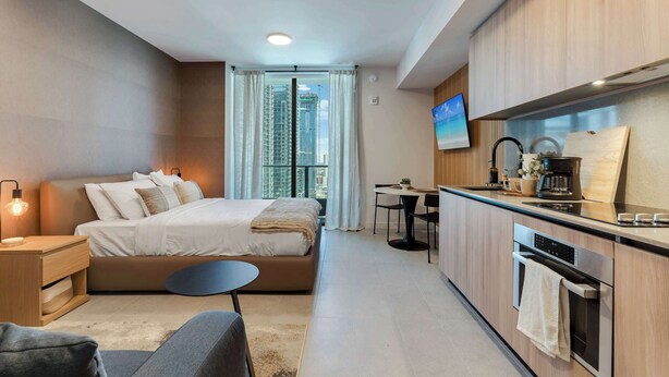 Ein modernes Hotelzimmer mit Bett, Nachttisch, Sessel, Küchenzeile und Fernseher.