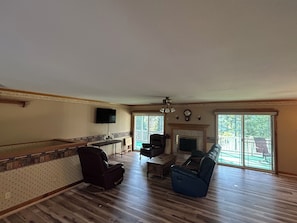 A spacious living room with hardwood flooring, a fireplace, a television mounted on the wall, and a large window providing a view of the outdoors.