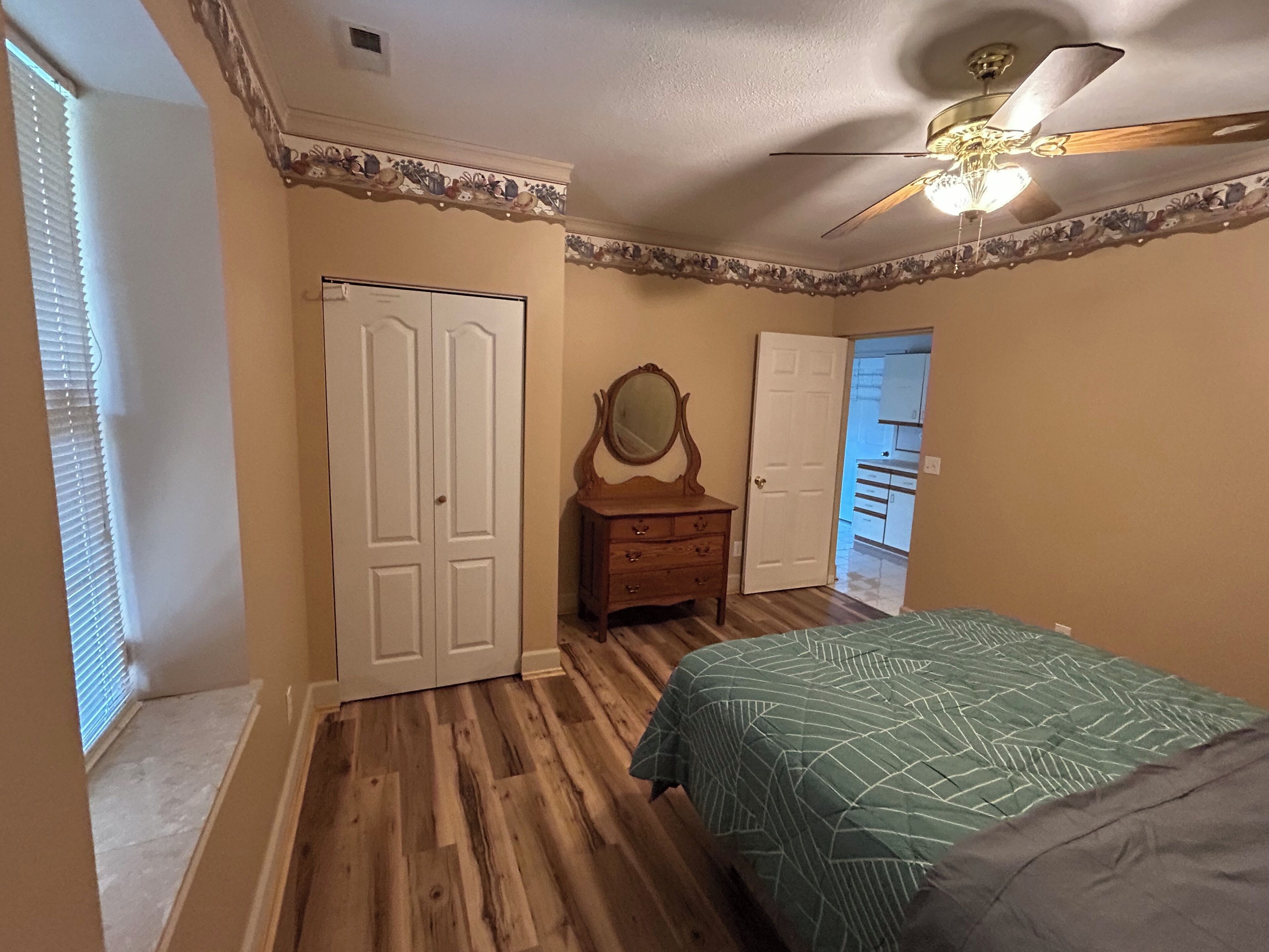 A bedroom with a wooden floor, a bed with a patterned bedspread, a dresser with a mirror, and a ceiling fan.