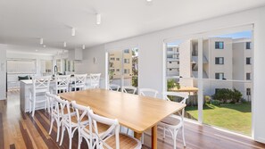 En rummelig, moderne lejlighed med et stort spiseområde, hvidt møblement og store vinduer med udsigt over de omkringliggende bygninger og grønne områder.