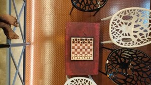 A chessboard on a table with a patterned tablecloth, surrounded by four chairs with intricate designs.