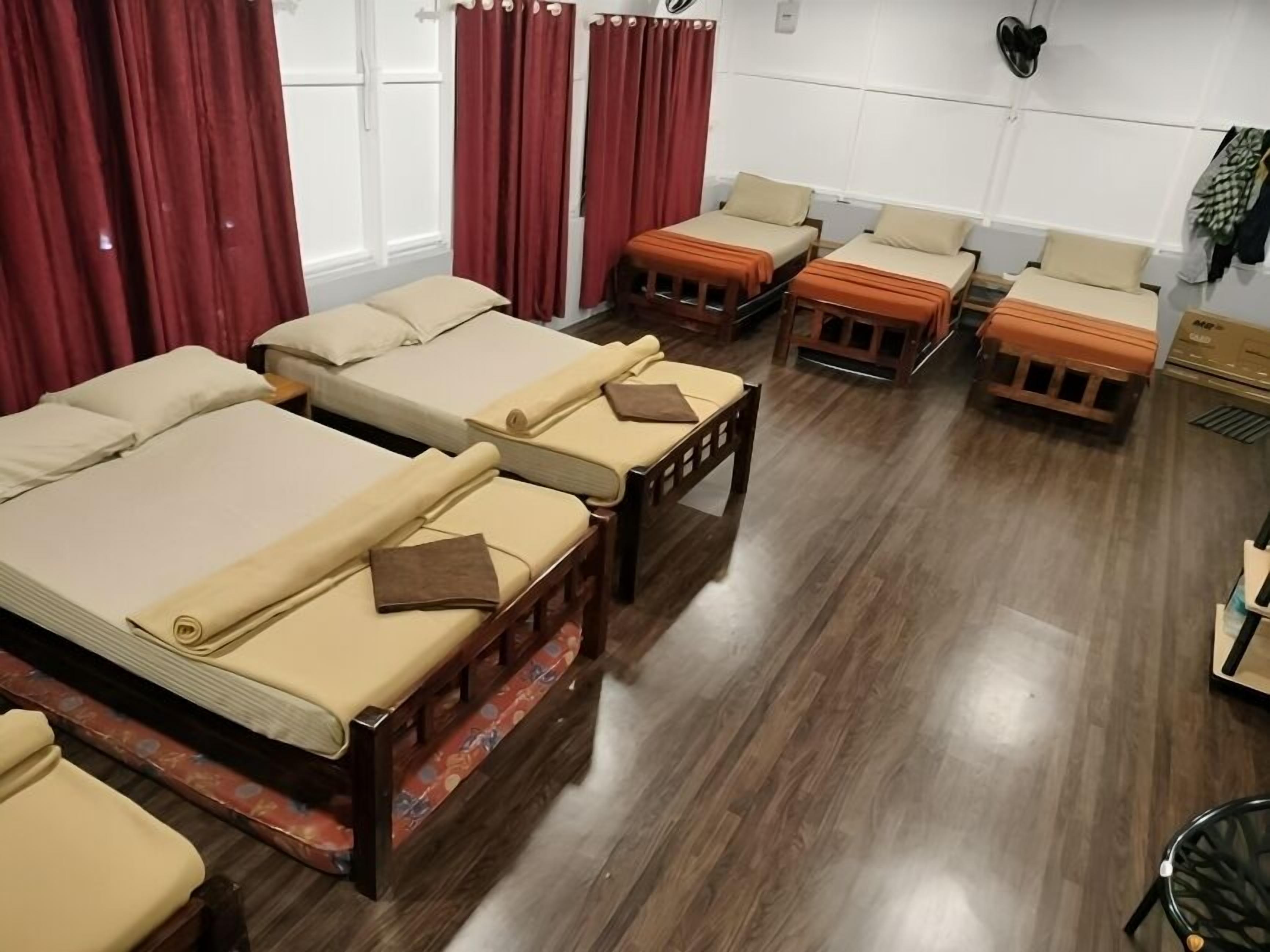 A room with multiple beds arranged in a row, wooden flooring, and a window with curtains.