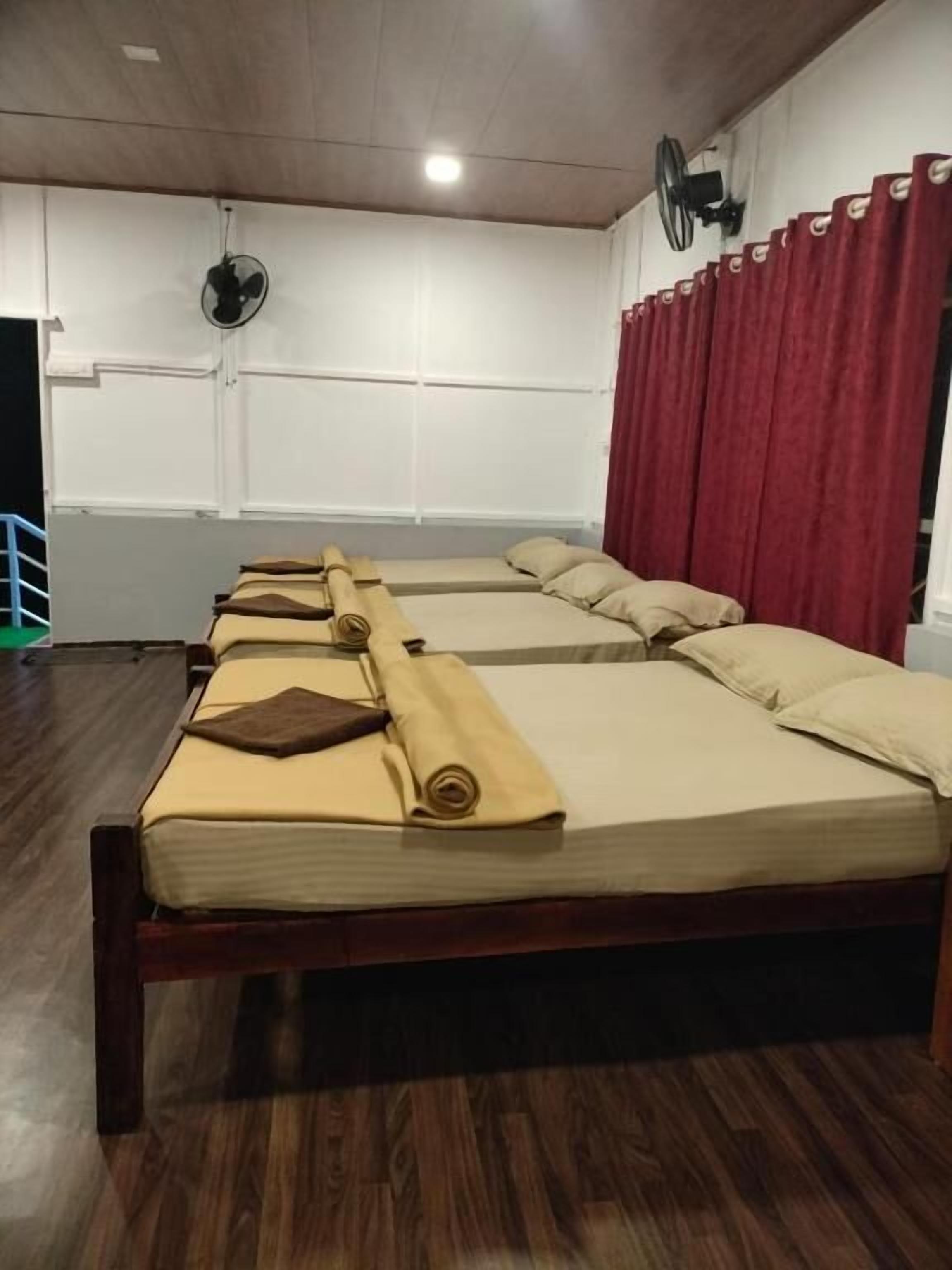 Une pièce avec quatre lits alignés en rangée, un plancher en bois, un rideau rouge et un ventilateur de plafond.