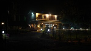 A two-story building with a balcony, illuminated by exterior lights, surrounded by a fence and vegetation.