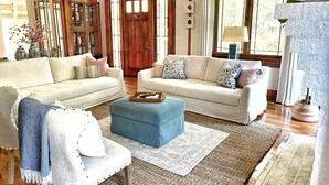 A spacious living room with two sofas, a blue ottoman, and a large window. - Pristine Craftsman 1900s Bungalow w Pool & Hot Tub (Savannah)