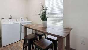 Una cocina funcional con una mesa de madera y taburetes, lavadora y una planta en maceta junto a la ventana.