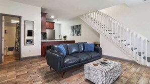 Una sala de estar con un sofá de cuero negro, un reposapiés estampado, un área de cocina y una escalera blanca.