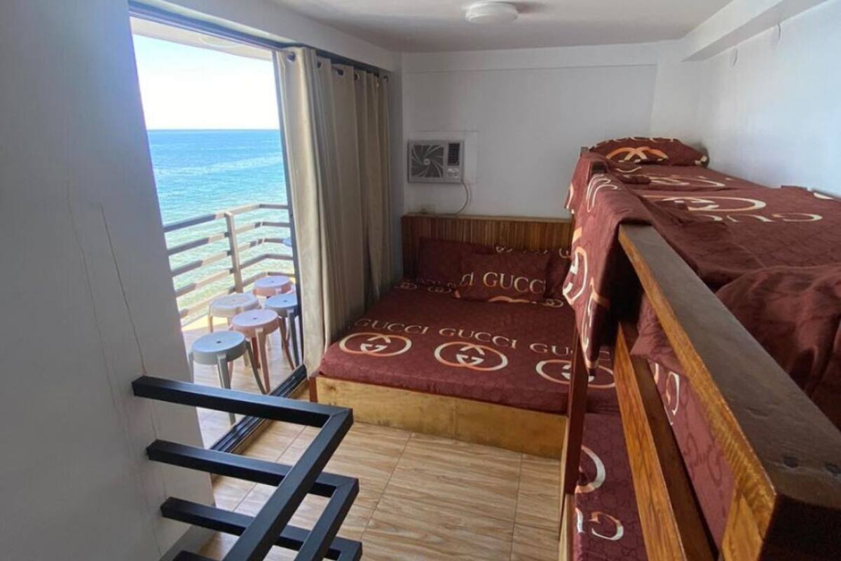 A balcony with a view of the sea, a bed with a Gucci-themed bedspread, a small table, and a ceiling fan.