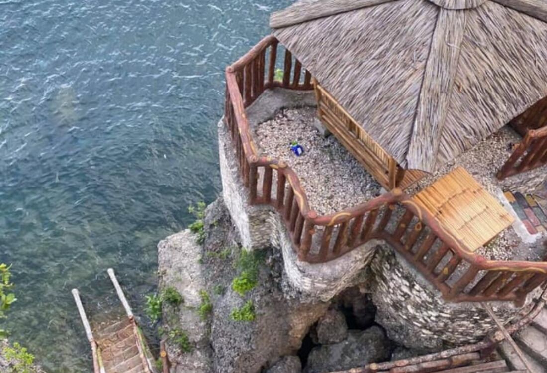 Une structure en bois dotée d’un toit de chaume, d’une terrasse en bois et d’une balustrade donnant sur une étendue d’eau.