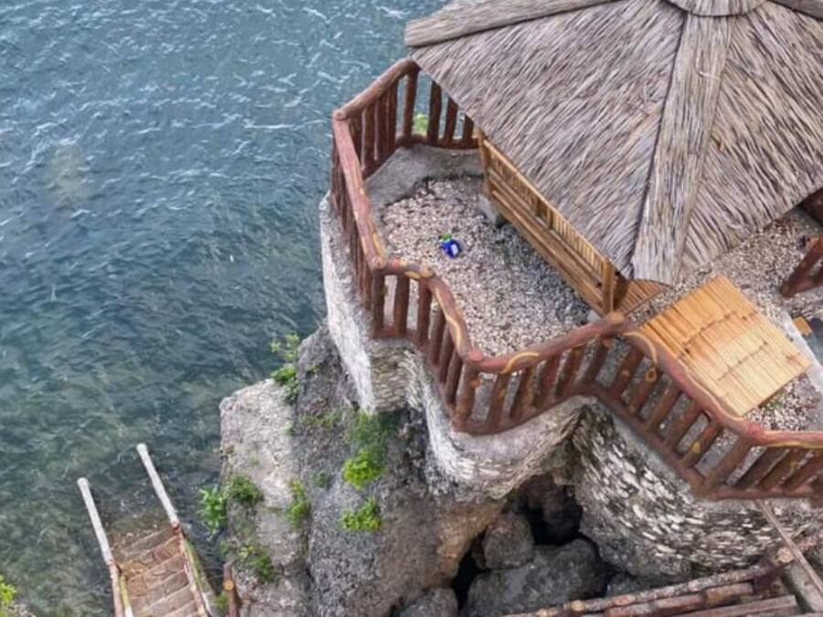 Eine Holzhütte mit einem Reetdach, einer Holzterrasse und einem Geländer mit Blick auf ein Gewässer.