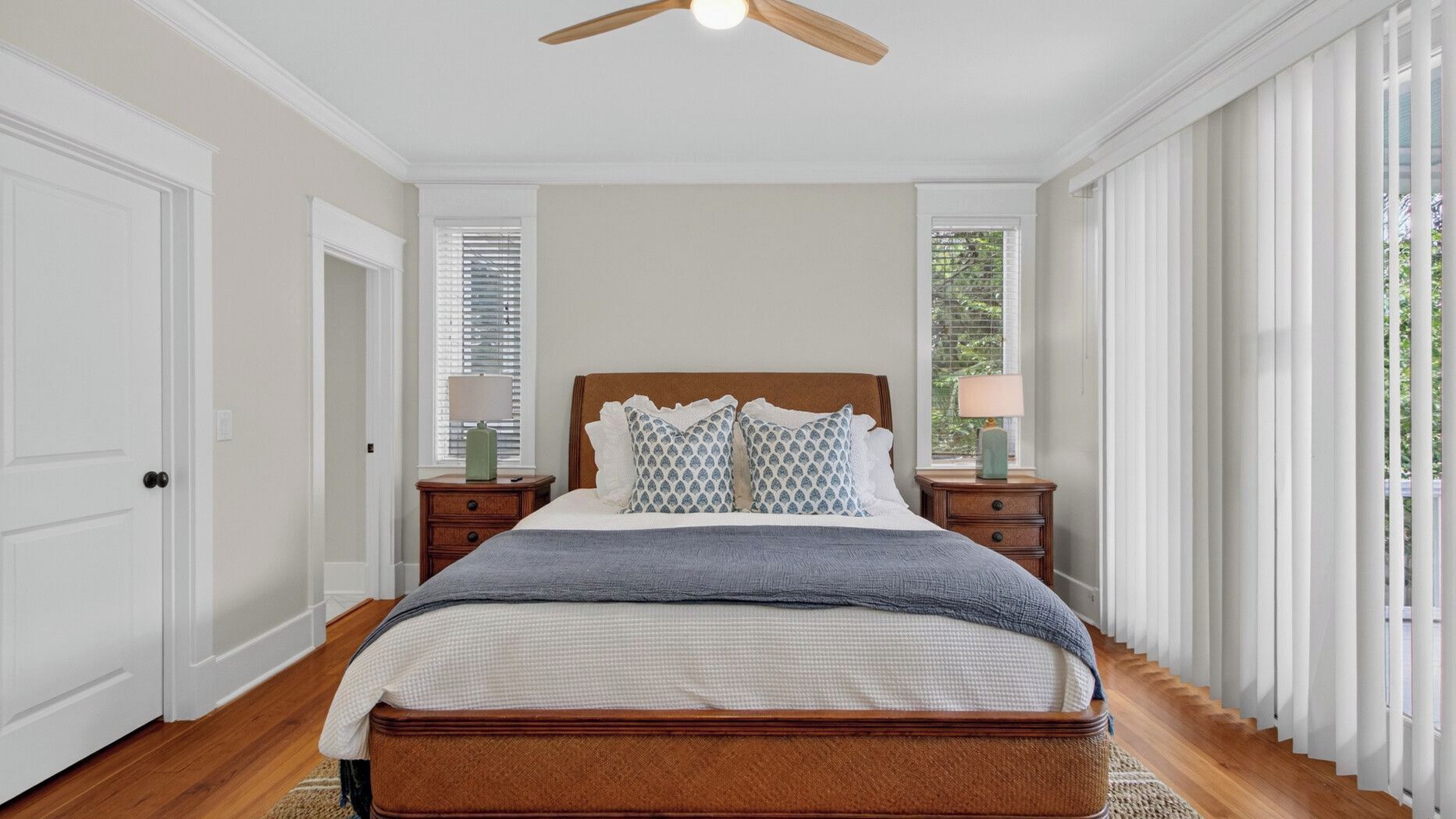 A bedroom with a wooden bed, two bedside tables with lamps, a ceiling fan, and large windows with blinds.