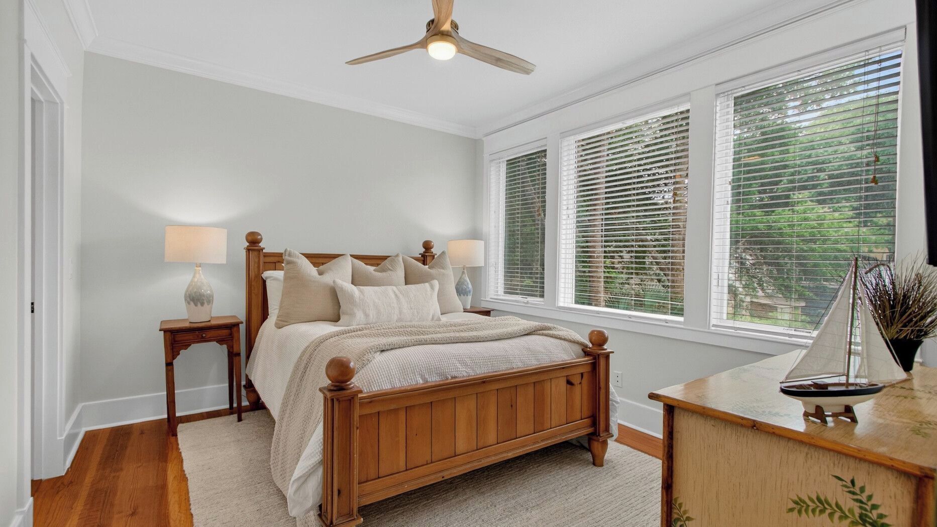 A bedroom with a wooden bed, bedside lamps, a nightstand, a desk, a sailboat decoration, and a window with blinds.
