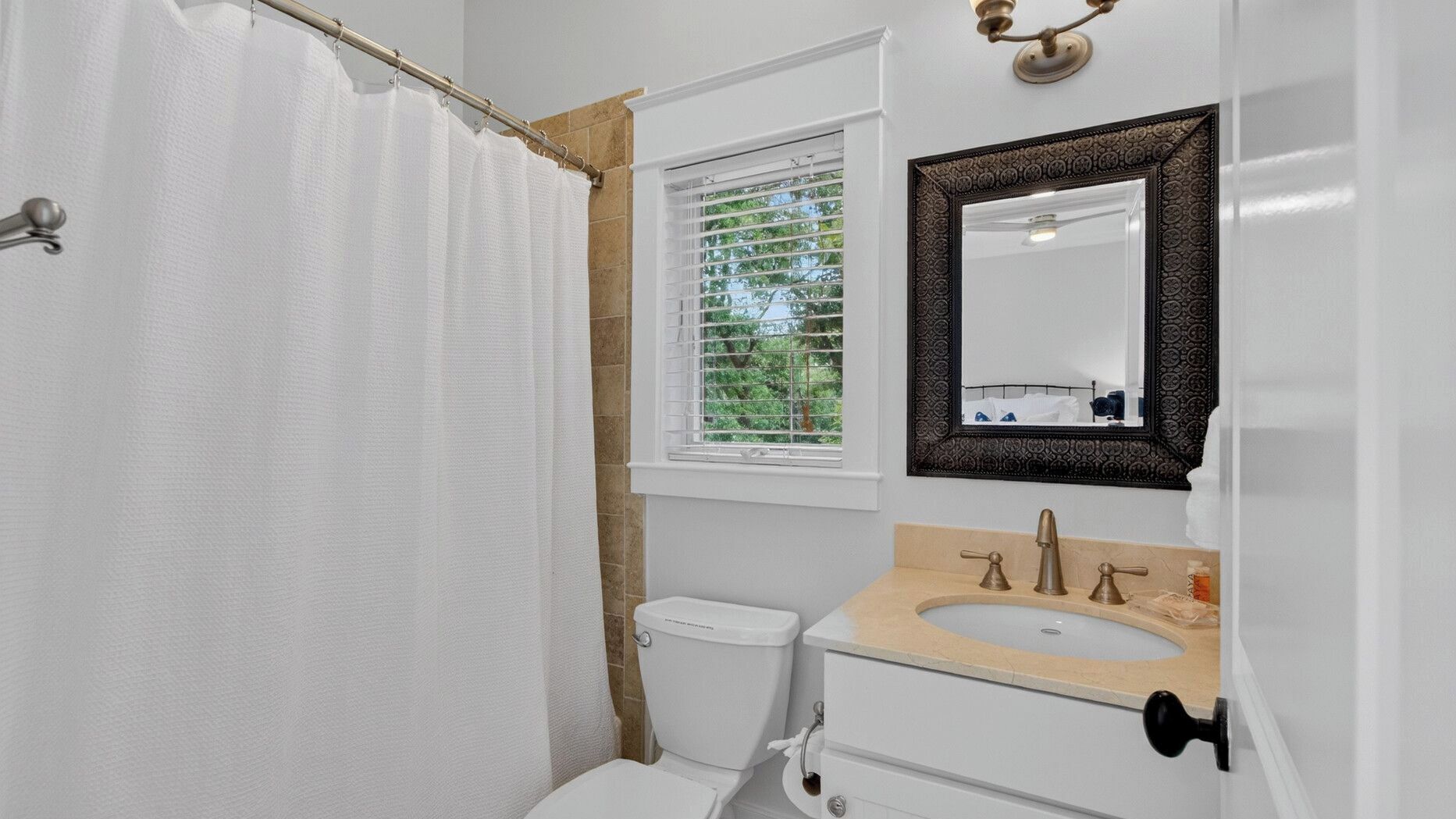 A bathroom with a shower curtain, a window with blinds, a toilet, a sink with a mirror above it, and a towel rack.