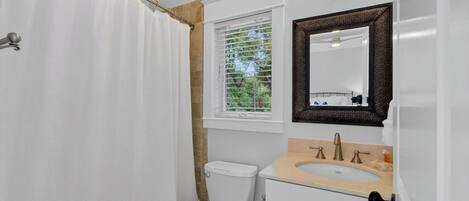 A bathroom with a shower curtain, a window with blinds, a toilet, a sink with a mirror above it, and a towel rack.