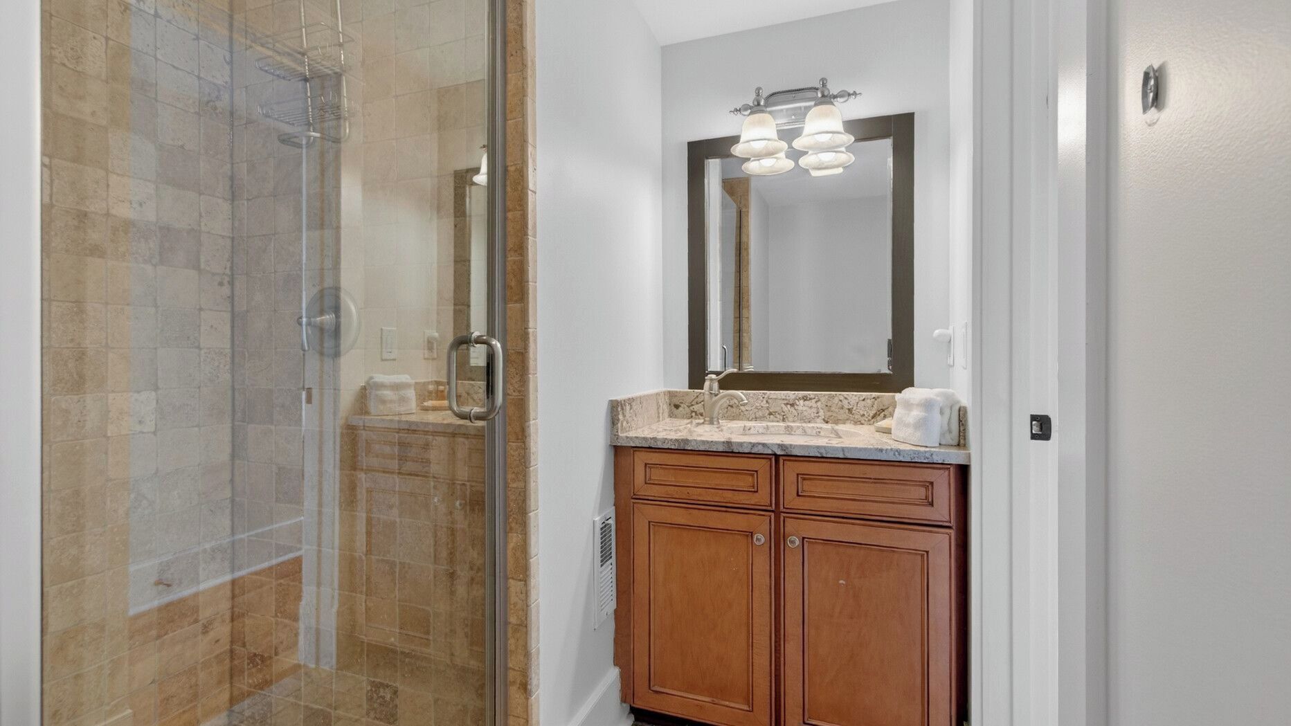 A bathroom with a glass shower enclosure, a sink with a marble countertop, and wooden cabinets.