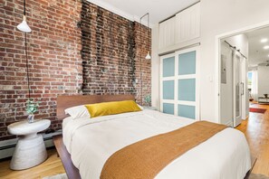 Ein Schlafzimmer mit Backsteinwand, einem Bett mit weißer Bettwäsche, einem Holz-Kopfteil und einer weißen Tür mit sandgestrahlten Glaselementen.