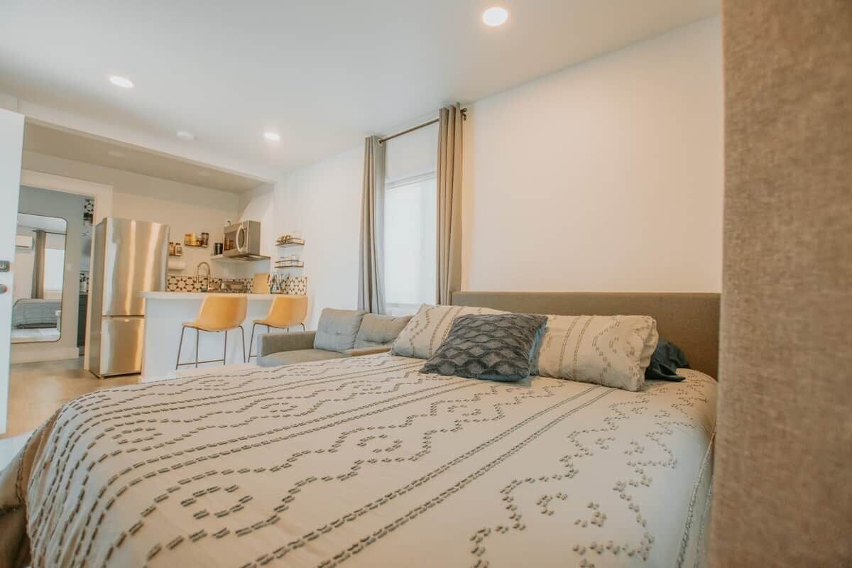 A bedroom with a bed, a desk, chairs, and a kitchenette.