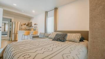 A bedroom with a bed, a desk, chairs, and a kitchenette.