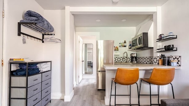A modern kitchen with a refrigerator, microwave, and coffee maker. There are two bar stools and a hanging rack with clothes. - ABQ Stunner Studio! Cute kitchen! Private parking! (Albuquerque)