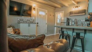 A living room with a leather couch, a TV mounted on the wall, and a kitchen area with a counter and stools. - ABQ Hub Studio - Old Town Gem! (Albuquerque)