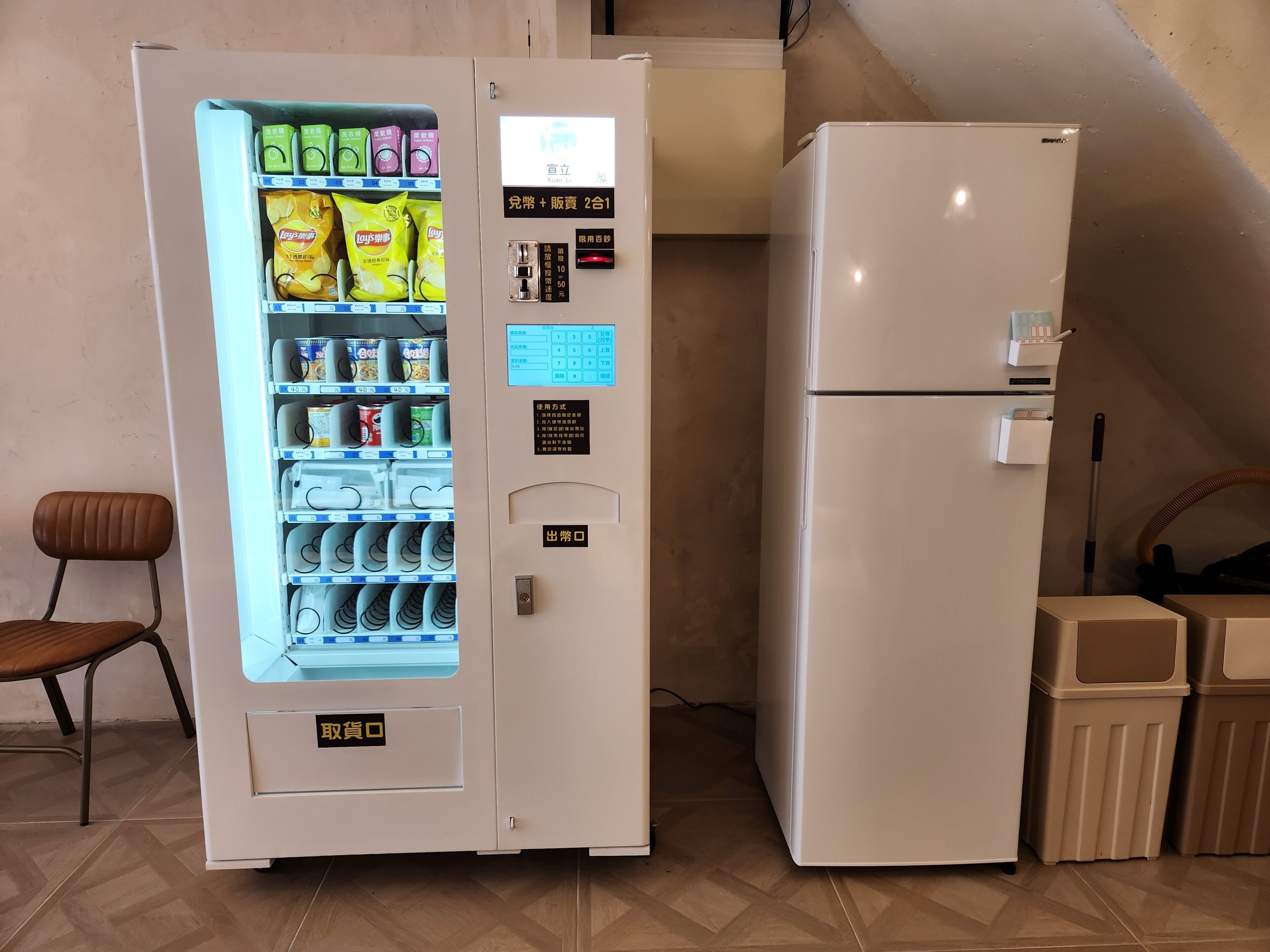 A vending machine with snacks and drinks, a refrigerator, a chair, and trash bins.