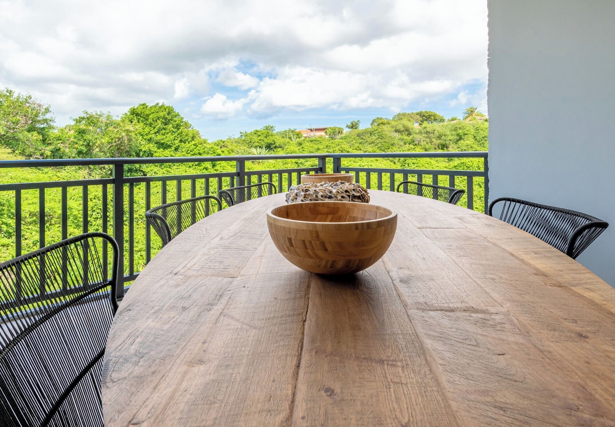 A wooden table with chairs on a balcony with a view of greenery and a building in the distance.