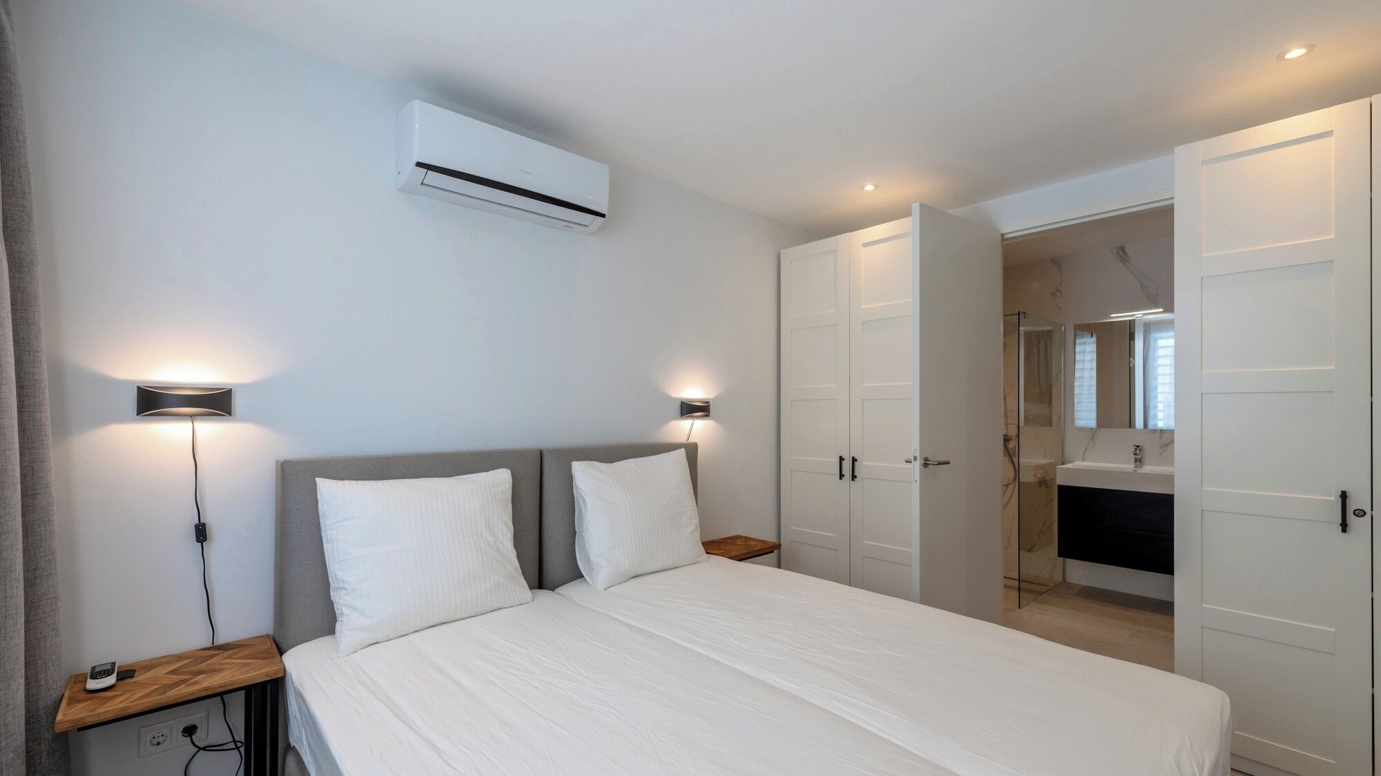 A bedroom with a bed, bedside tables, wall-mounted lights, an air conditioner, and a bathroom with a shower and mirror.