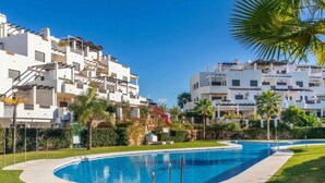 Una piscina rodeada de edificios blancos con balcones, palmeras y un cielo azul despejado.