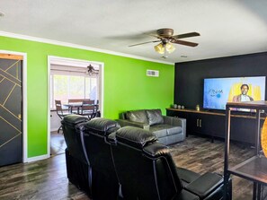 Ein Wohnzimmer mit grüner Wand, einem schwarzen Ledersofa, einem Deckenventilator und einem Fernseher, der einen Film zeigt.
