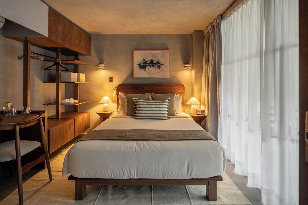 A hotel room with a bed, bedside tables, a desk, a chair, and a hanging coat rack.