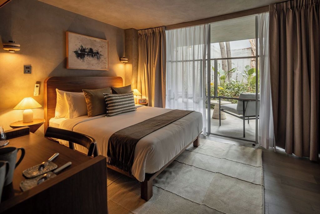A hotel room with a bed, bedside tables, a desk, a chair, a balcony with a table and chairs, and a view of greenery.