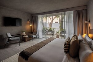 Una habitación de hotel moderna con una cama grande, un televisor montado en la pared, un área de descanso con sillas y una mesa, y vistas amplias a través de grandes ventanales.