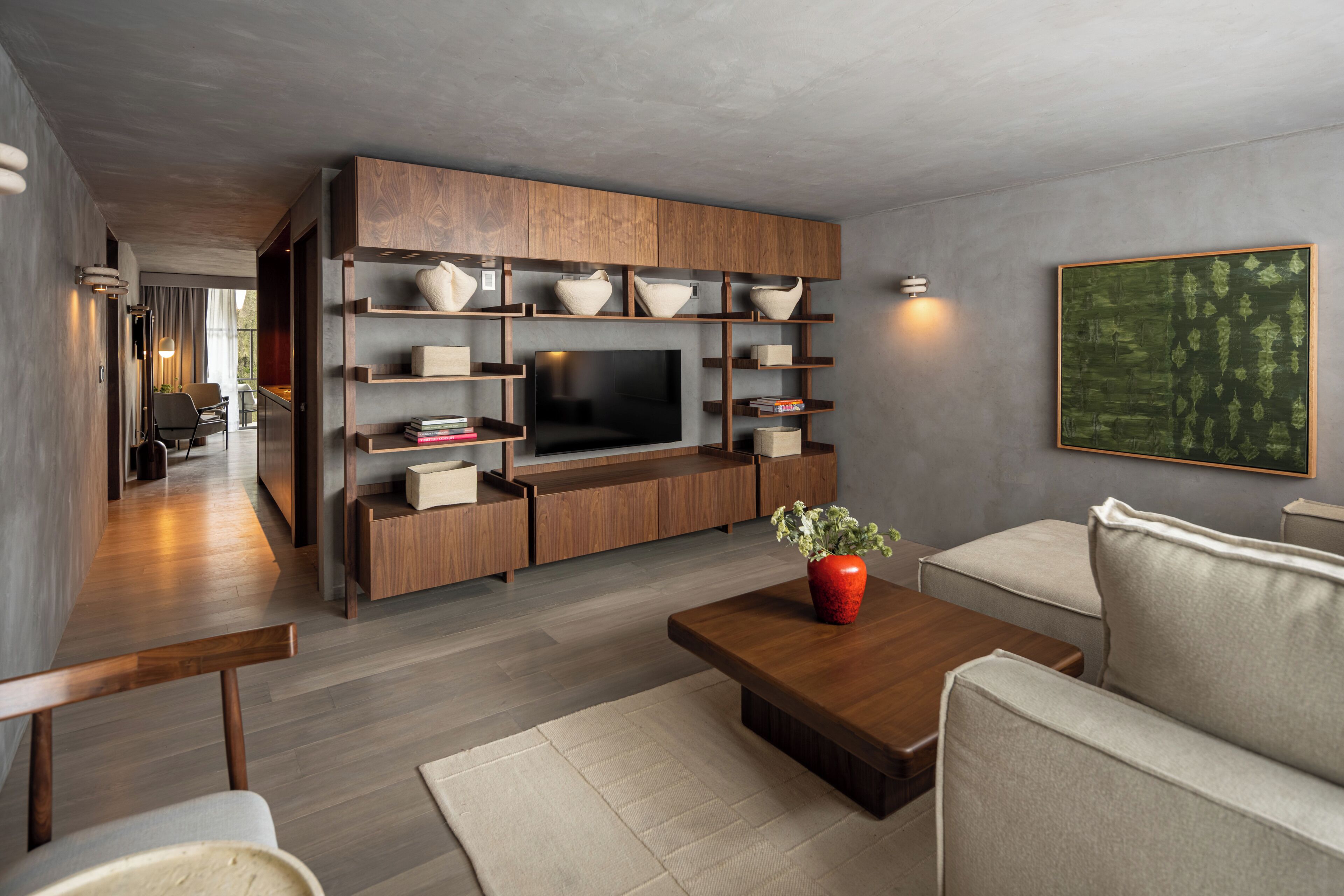 A modern living room with a flat-screen TV, wooden shelving, a red vase, and a beige sofa.
