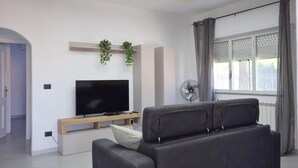 A modern living room with a flat-screen TV, a sofa, a small wooden table, and a window with curtains. - 3 bedroom stunning home in Trabia (Trabia)