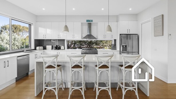 Una cucina moderna con mobili bianchi, un'isola centrale con sgabelli da bar, un frigorifero in acciaio inossidabile e vista sugli alberi.