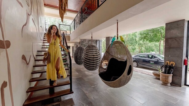 A woman descending a wooden staircase in a modern interior with hanging chairs and a parked car visible. - Talulah Living 3BR Apt - Private & Shared Pool (Tulum)