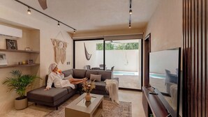 A modern living room with a sofa, a coffee table, a TV, and a large window. - Talulah Living 3BR Apt - Private & Shared Pool (Tulum)