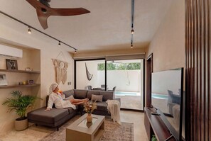 A modern living room with a sofa, a coffee table, a TV, and a large window. - Talulah Living 3BR Apt - Private & Shared Pool (Tulum)
