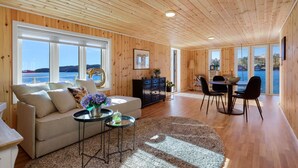 Een gezellige woonkamer met een bank, salontafel en eethoek, grote ramen met uitzicht op de zee, en houten lambrisering aan de muren en het plafond.