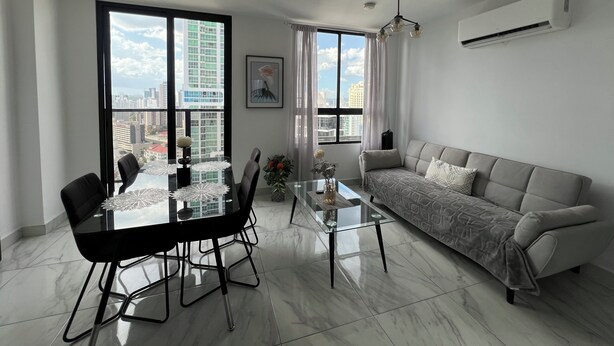 Ein modernes Wohnzimmer mit einem Glastisch, schwarzen Stühlen, einem grauen Sofa und Stadtblick durch große Fenster.
