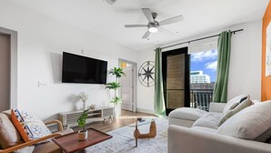 Ein modernes Wohnzimmer mit Flachbildfernseher, Deckenventilator, großem Fenster mit Vorhängen und Blick auf die Stadt.